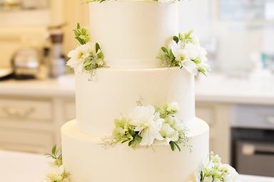 Three-tier wedding cake with fresh white flowers and delicate green leaves in a bright bakery setting